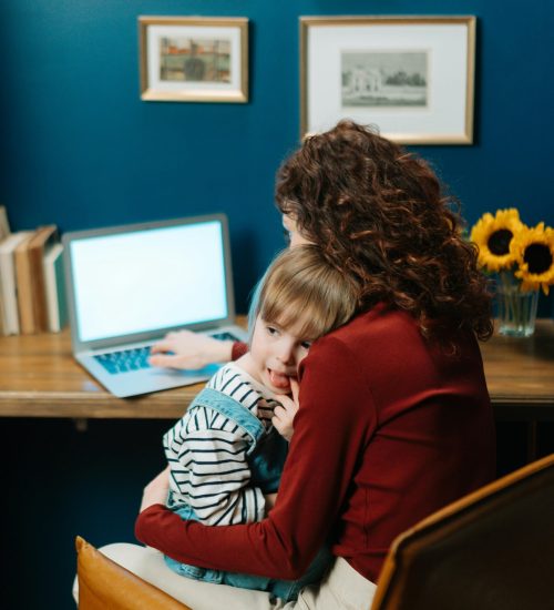 pexels-photo-6985723-6985723 A mother works on a laptop with her child sitting on her lap, showcasing work-life balance.
